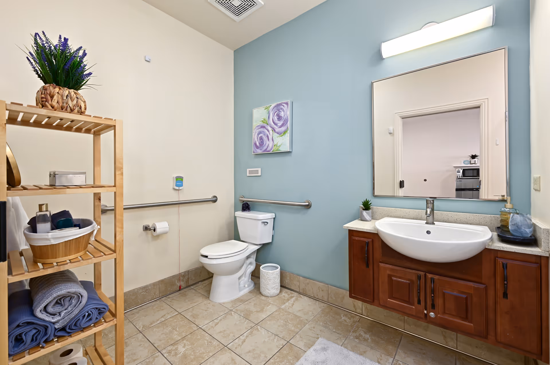 A clean and spacious bathroom with beige and light blue walls. The bathroom features a white toilet with grab bars on the walls, a wooden shelving unit with towels and toiletries, a large mirror above a wooden vanity with a white sink, and a small potted plant on the counter. There is a painting of purple flowers on the blue wall.