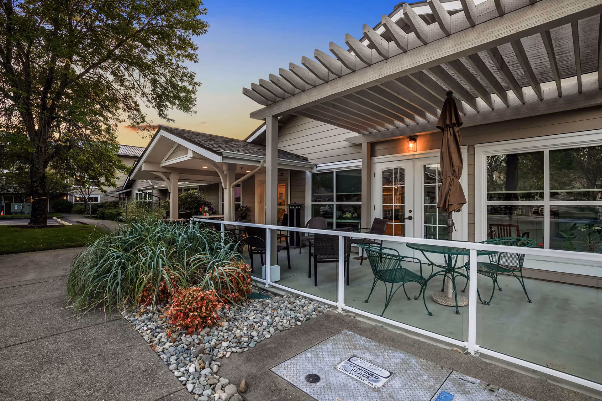 Exterior patio of a senior living building with a pergola, outdoor tables and chairs, and landscaping at dusk.