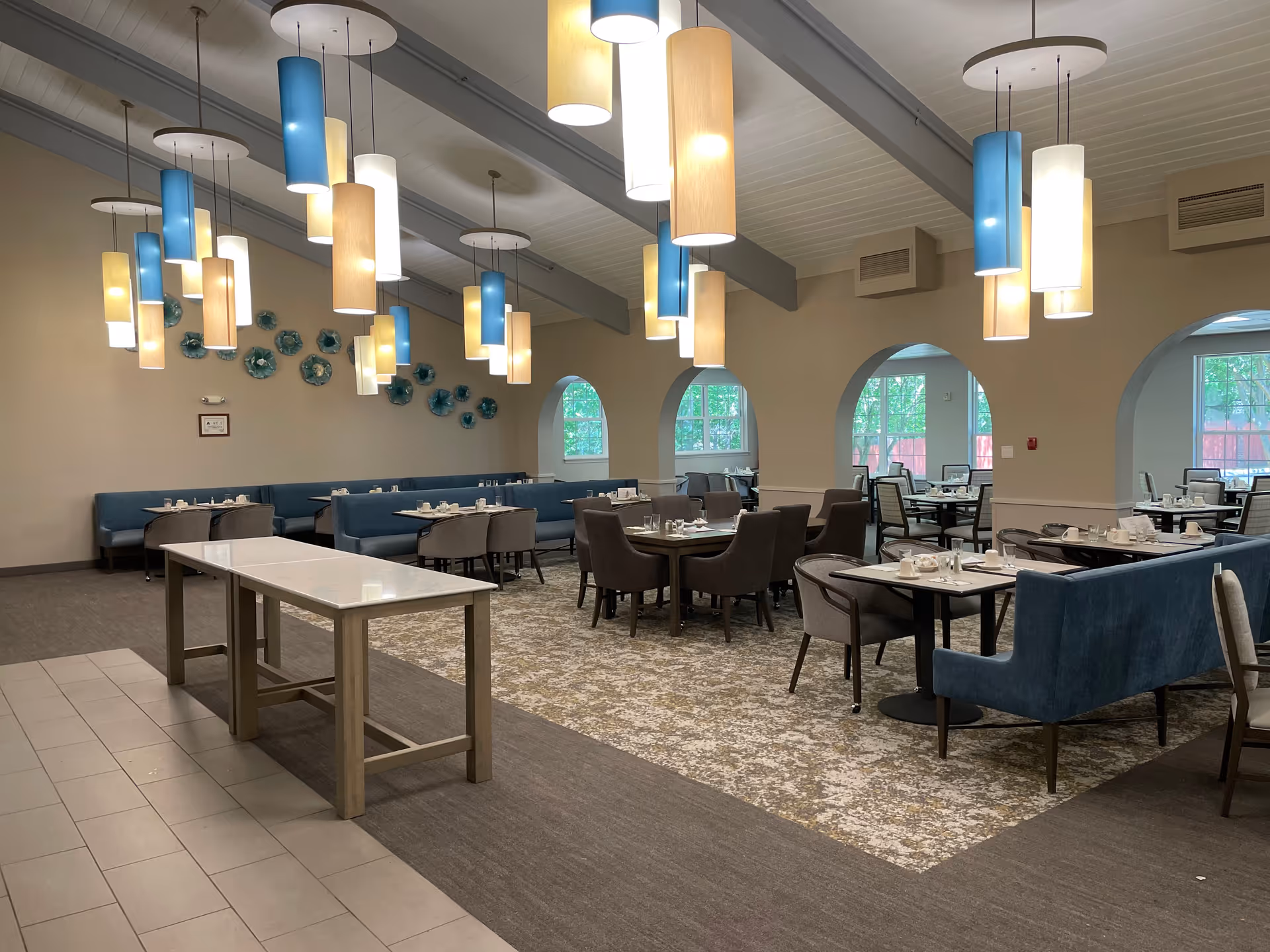 Large dining room with hanging cylindrical pendant lights, blue upholstered booths and multiple tables set for service.