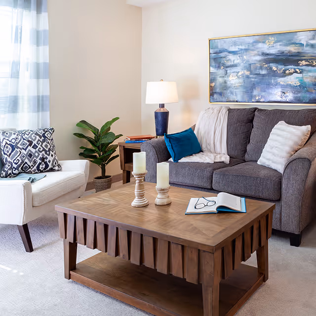 A cozy living room with a gray sofa adorned with white and blue pillows, a wooden coffee table with two candles and an open book with glasses on it, a white armchair with a patterned pillow, a potted plant, a side table with a lamp and books, and a large abstract painting on the wall.
