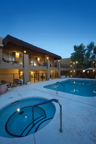 Outdoor view of Scottsdale Village Square at dusk featuring a swimming pool and a hot tub surrounded by a two-story building with balconies and outdoor seating areas.