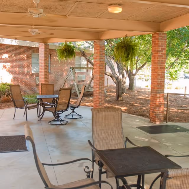 Covered outdoor patio with tables and chairs, brick columns, hanging ferns and nearby trees.