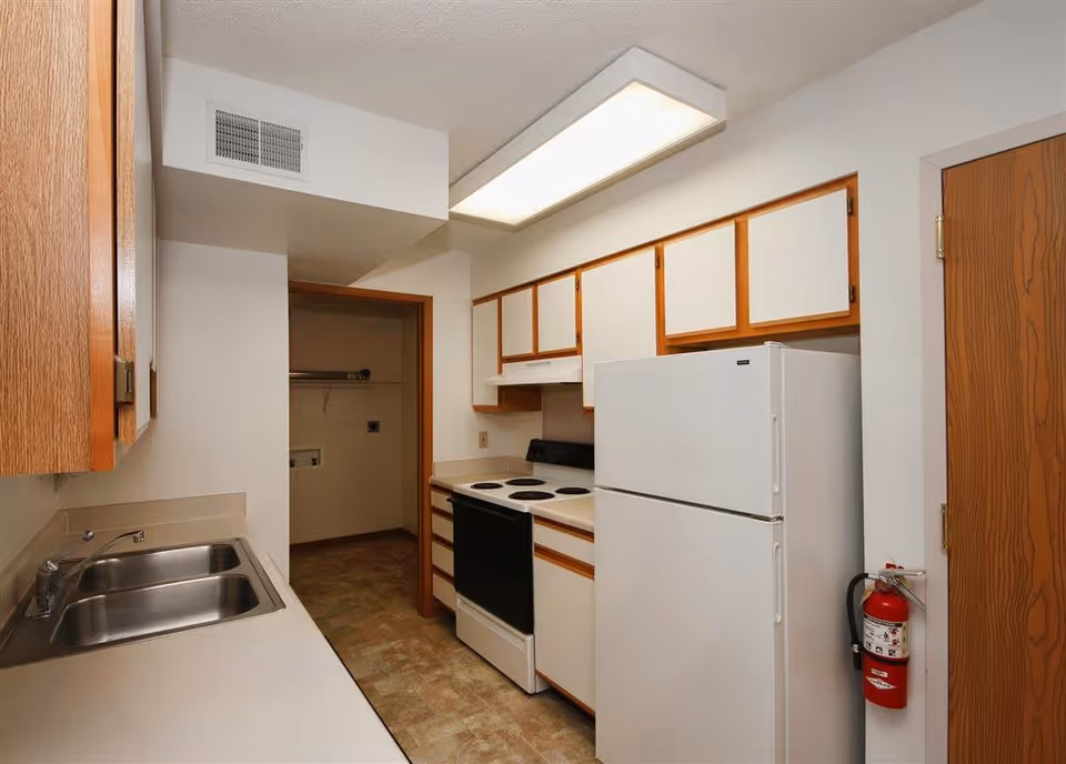 A small kitchen with white countertops, a double stainless steel sink, white cabinets with wooden trim, a white refrigerator, and a white electric stove with four burners. There is a fire extinguisher mounted on the wall near a wooden door, and a doorway leading to a small pantry or laundry area. The ceiling has a rectangular fluorescent light fixture.