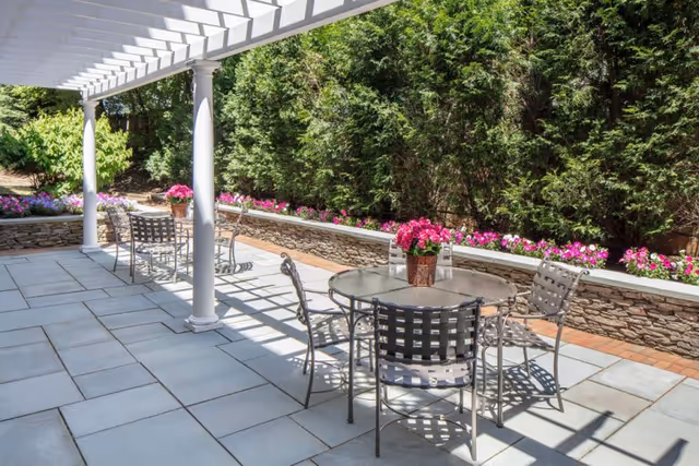 Outdoor patio area with a white pergola overhead, metal tables and chairs, stone tiled floor, and a stone retaining wall with pink and white flowers and tall green bushes in the background.