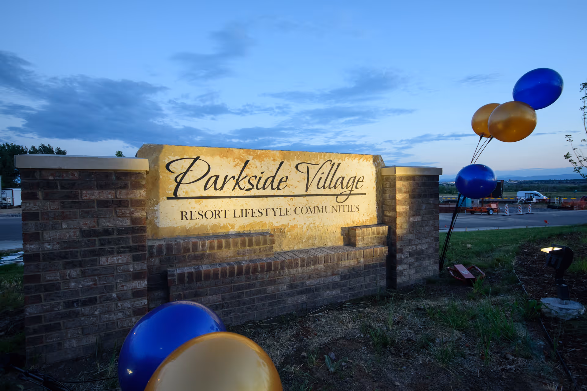 Illuminated stone and brick sign for Parkside Village Resort Lifestyle Communities at dusk, with blue and gold balloons tied to a post nearby and a partly cloudy sky in the background.