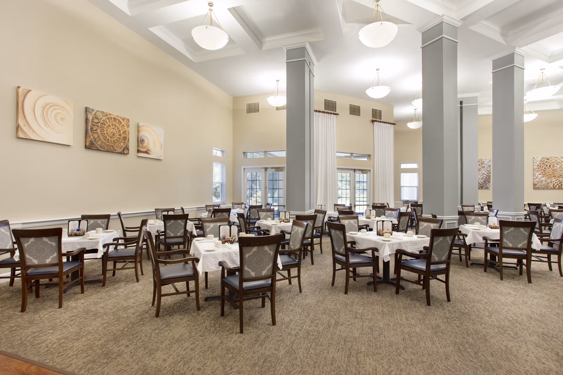A spacious dining room in a senior living facility with multiple tables covered in white tablecloths, each set with cups, plates, and condiments. The room features tall ceilings with hanging light fixtures, large windows with white curtains, and decorative wall art depicting seashells. Dark wooden chairs with patterned upholstery surround the tables.