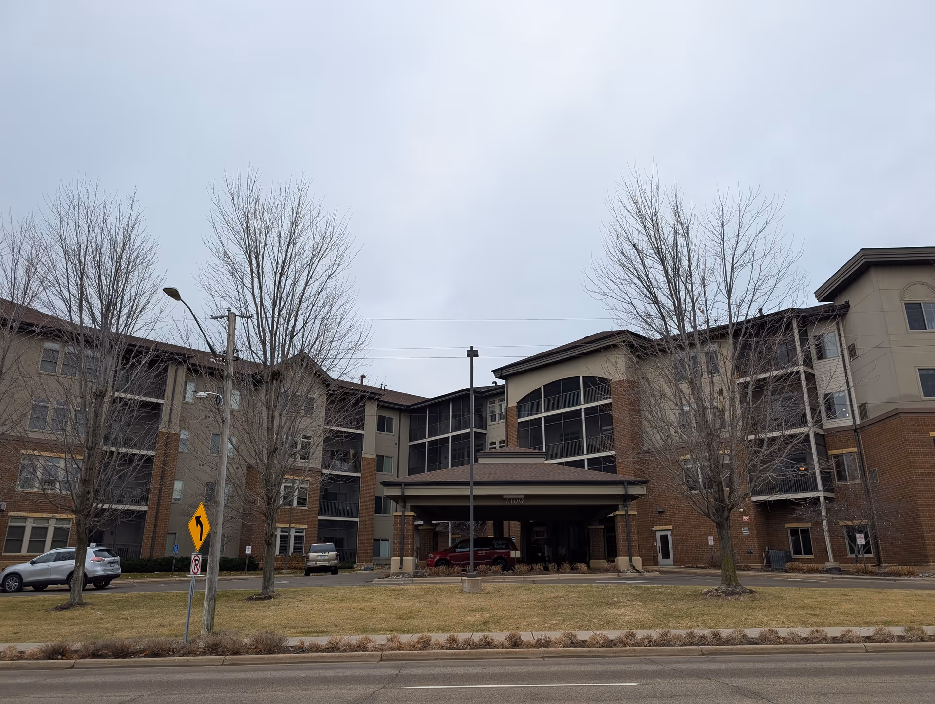 Exterior view of a multi-story senior living facility building with a covered entrance and several leafless trees in front. There are a few parked cars and a road with traffic signs in the foreground under an overcast sky.