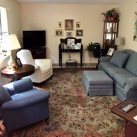 A cozy living room with a patterned area rug, a blue sofa with matching ottomans, a white armchair, and a blue armchair. There is a wooden side table with a lamp and framed photo, a TV on a stand in the corner, and a small table against the wall displaying framed pictures. A glass-front cabinet with flowers on top and a floor lamp are also visible.