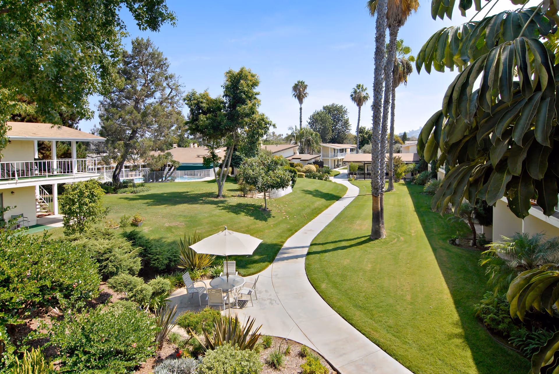 A sunny outdoor garden area at Grossmont Gardens Senior Living featuring a winding concrete pathway, green lawns, various trees including palm trees, and a small seating area with a table and umbrella surrounded by chairs.