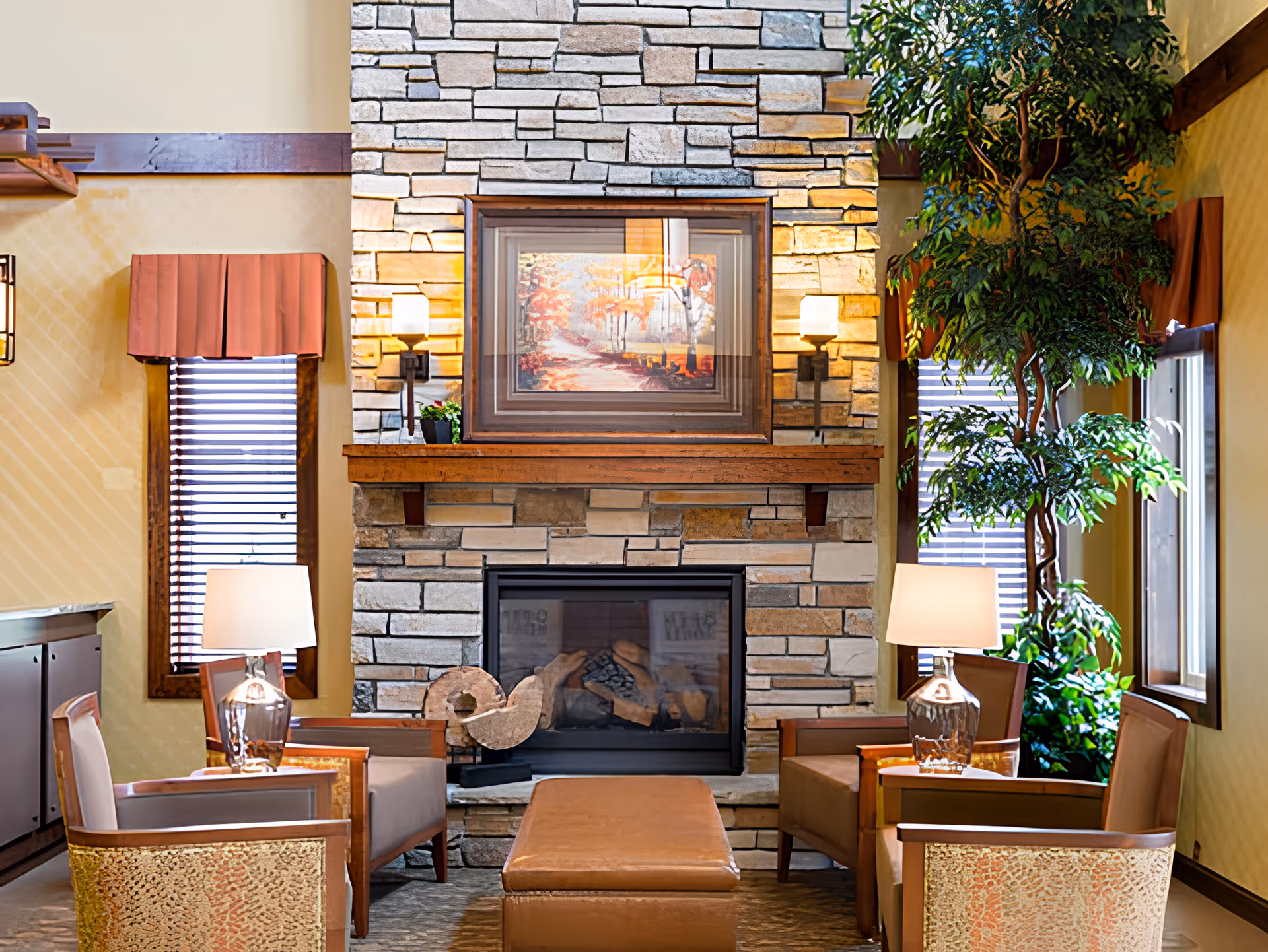 Cozy sitting area with a stone fireplace in the center, flanked by two windows with wooden blinds and red valances. Four armchairs surround a rectangular leather ottoman, with two table lamps on side tables next to the chairs. A large green plant is positioned near the right window, and a framed autumn landscape painting hangs above the fireplace.
