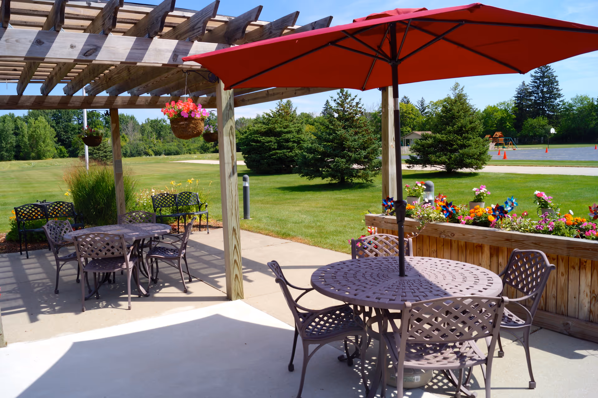 Outdoor patio area with metal tables and chairs, one table shaded by a large red umbrella. Wooden pergola overhead with hanging flower baskets. Green lawn and trees in the background under a clear blue sky.