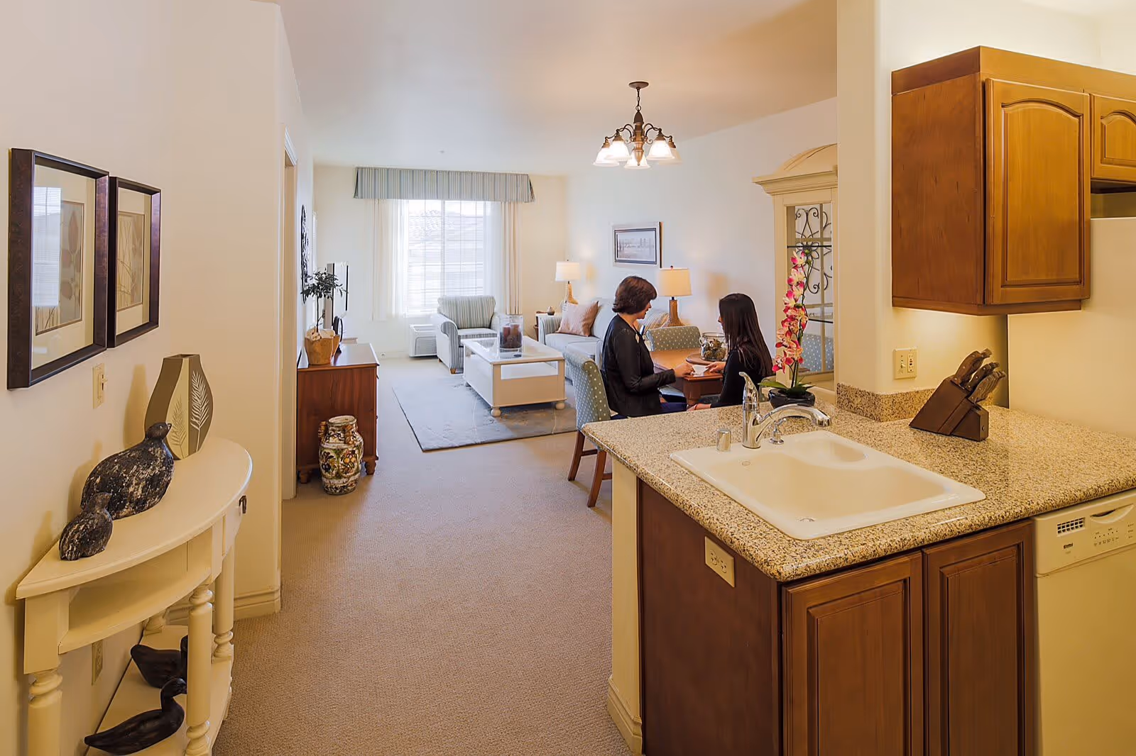 View of a senior living facility apartment showing a kitchen area with a sink and wooden cabinets in the foreground, and a living room with two women sitting and talking at a small dining table in the background. The living room has a sofa, armchair, coffee table, lamps, and a window with curtains letting in natural light.