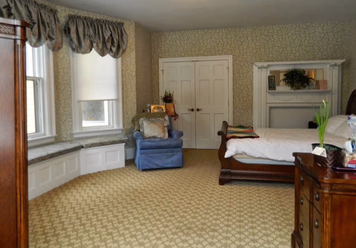 A bedroom with a large bed featuring a wooden frame and white bedding, a blue armchair with cushions, a wooden dresser with a plant and other items on top, two windows with valances and blinds, a double-door closet, and a decorative white fireplace mantel with a plant and framed pictures.
