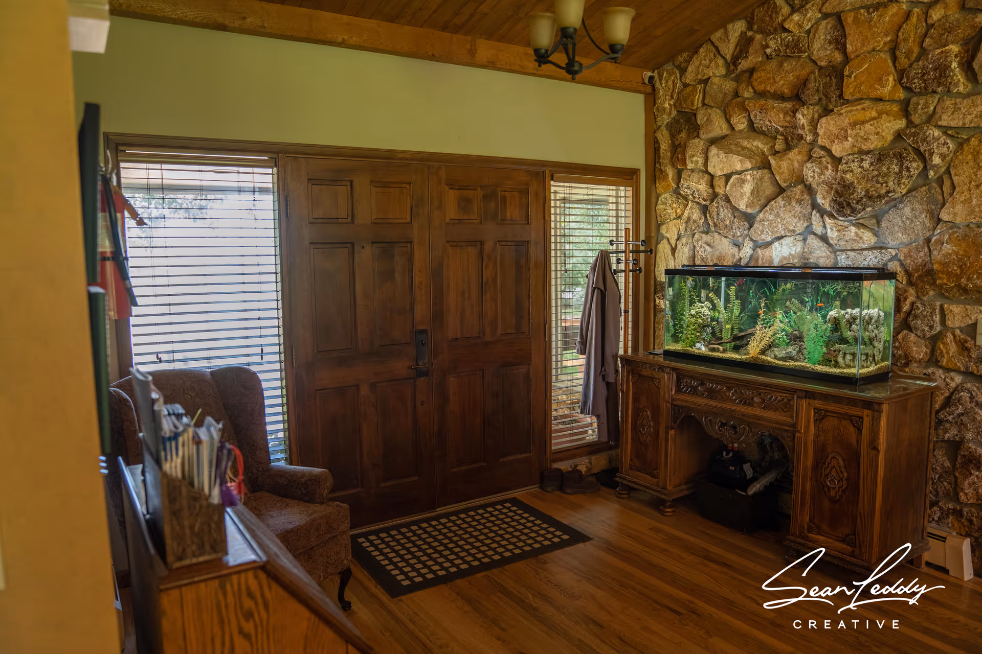 Interior view of an entryway with a wooden front door flanked by two windows with blinds. To the right, there is a stone wall with a wooden cabinet holding a large aquarium filled with plants and decorations. On the left side, there is a cushioned armchair and a wooden piece of furniture with magazines or papers organized on top. The floor is wooden, and a coat rack with a hanging garment is near the door.