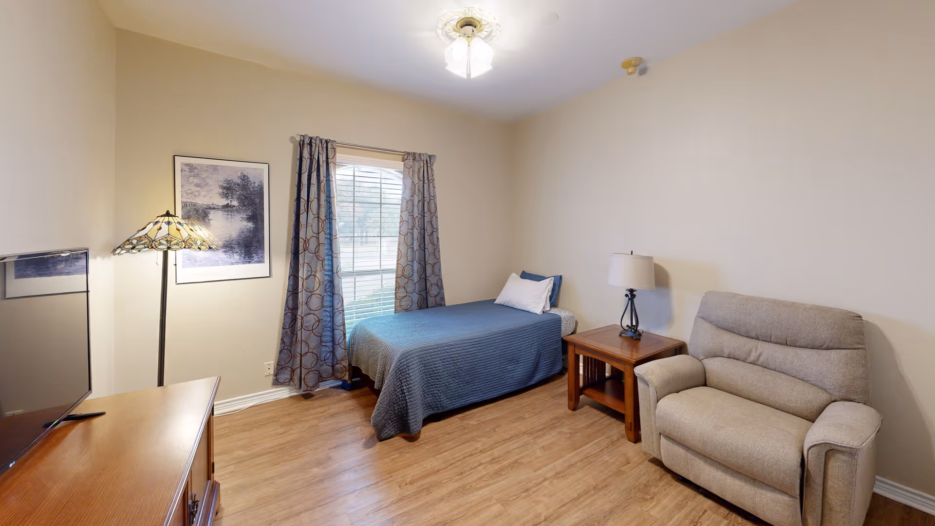 A senior living bedroom with a single bed covered in a blue quilt, a wooden nightstand with a lamp, a beige recliner chair, a floor lamp with a stained glass shade, a framed picture on the wall, a window with patterned curtains, and a wooden dresser with a TV on top.