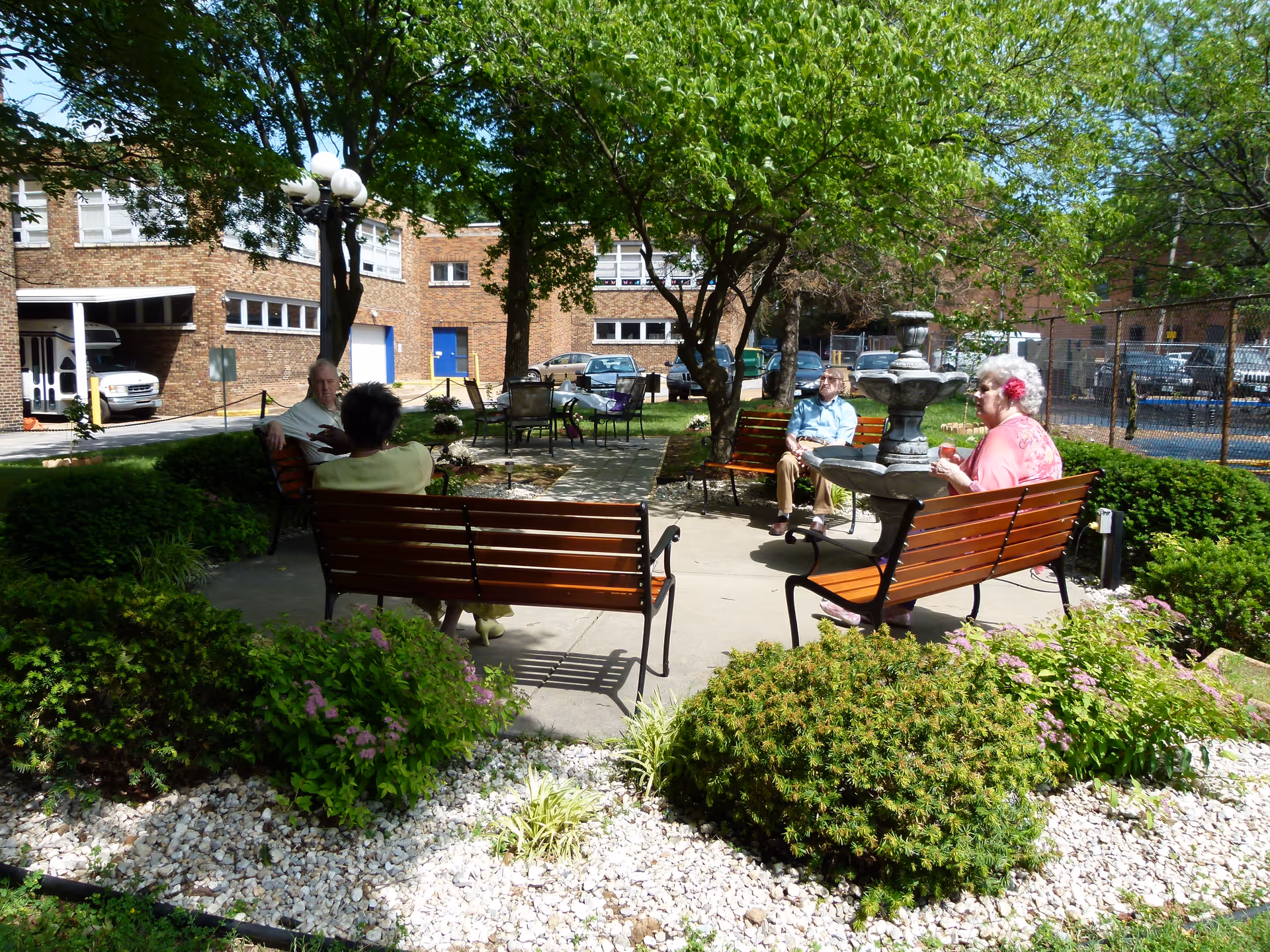 An outdoor garden area at Tower Grove Manor with several elderly people sitting on wooden benches around a concrete pathway. The garden features green bushes, flowering plants, a tree providing shade, and a multi-tiered stone fountain. In the background, there is a brick building and parked cars.