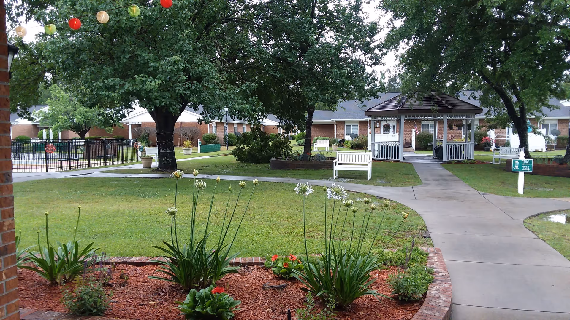 A well-maintained outdoor garden area at Truewood by Merrill, New Bern, featuring green grass, flower beds with blooming plants, several large trees, white benches, a gazebo, and paved walkways. There are red and white hanging lanterns visible in the upper left corner, and residential buildings in the background.