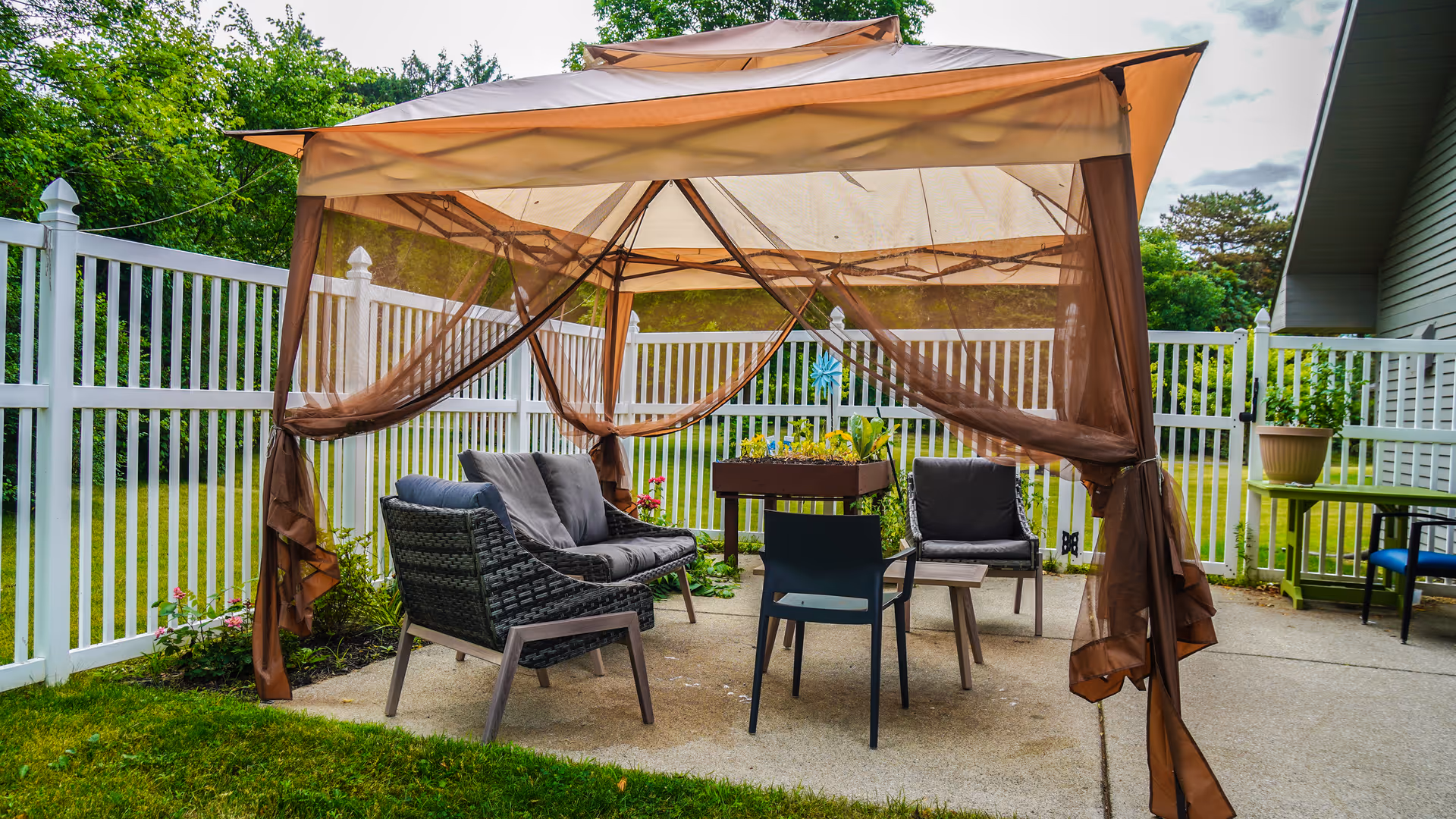 Outdoor patio area with a beige canopy tent featuring brown mesh curtains tied back. Under the canopy are cushioned wicker chairs, a black plastic chair, and a small wooden table. The area is enclosed by a white picket fence with green grass and trees in the background.