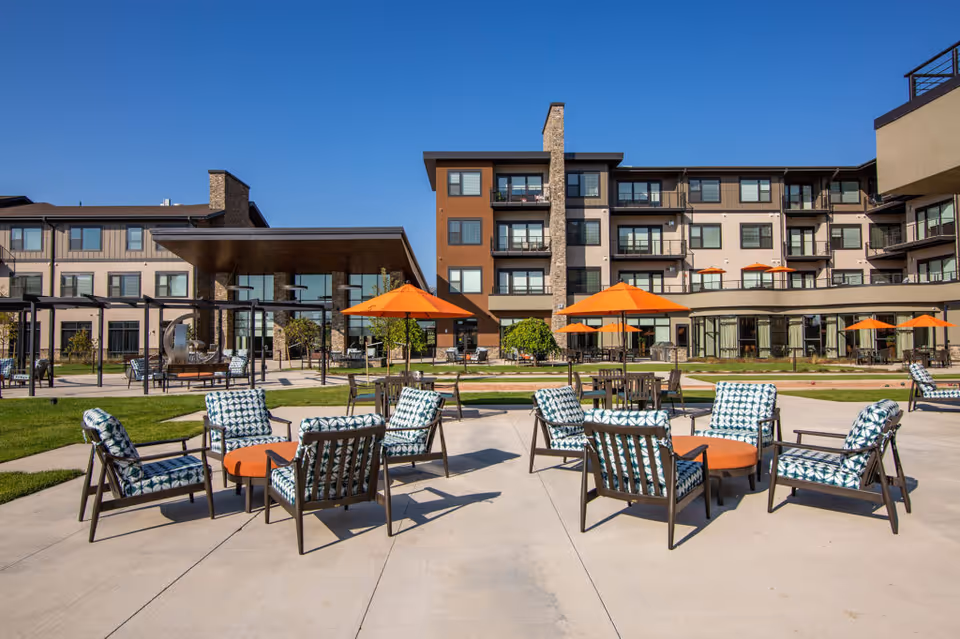 Outdoor seating area at The Ridge Pinehurst featuring cushioned chairs arranged around small tables with orange umbrellas, set on a concrete patio in front of a multi-story building under a clear blue sky.