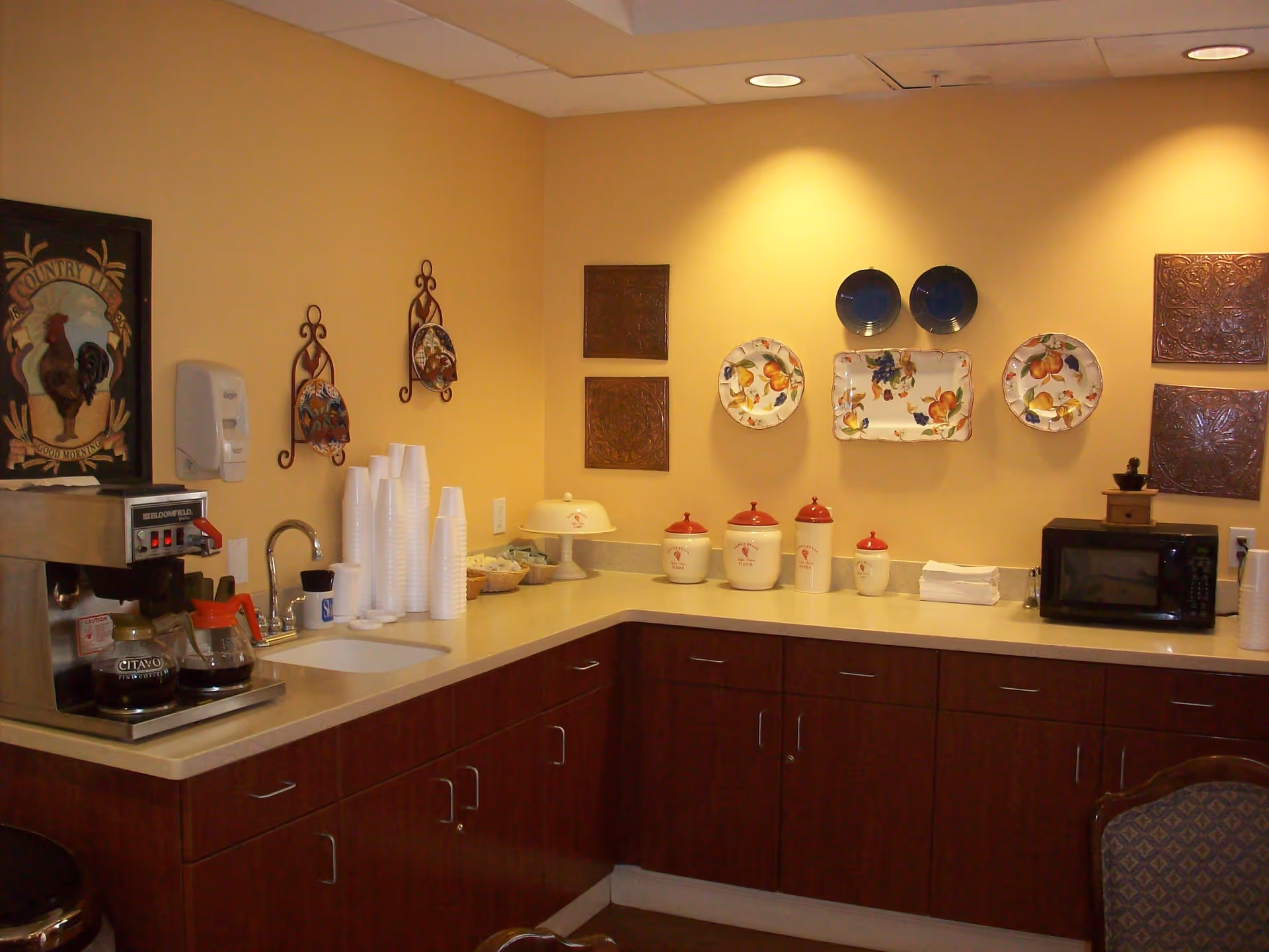 A small kitchen area with a coffee maker, stacks of disposable cups, a sink, decorative plates and wall hangings, a microwave, and several ceramic canisters on the countertop.