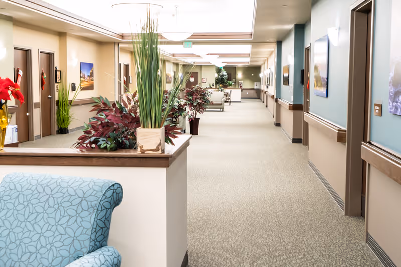A well-lit, clean hallway in a senior living facility with beige carpet and light-colored walls. The hallway is decorated with plants, framed pictures, and holiday stockings hanging on the doors. There is a blue patterned armchair partially visible in the foreground.