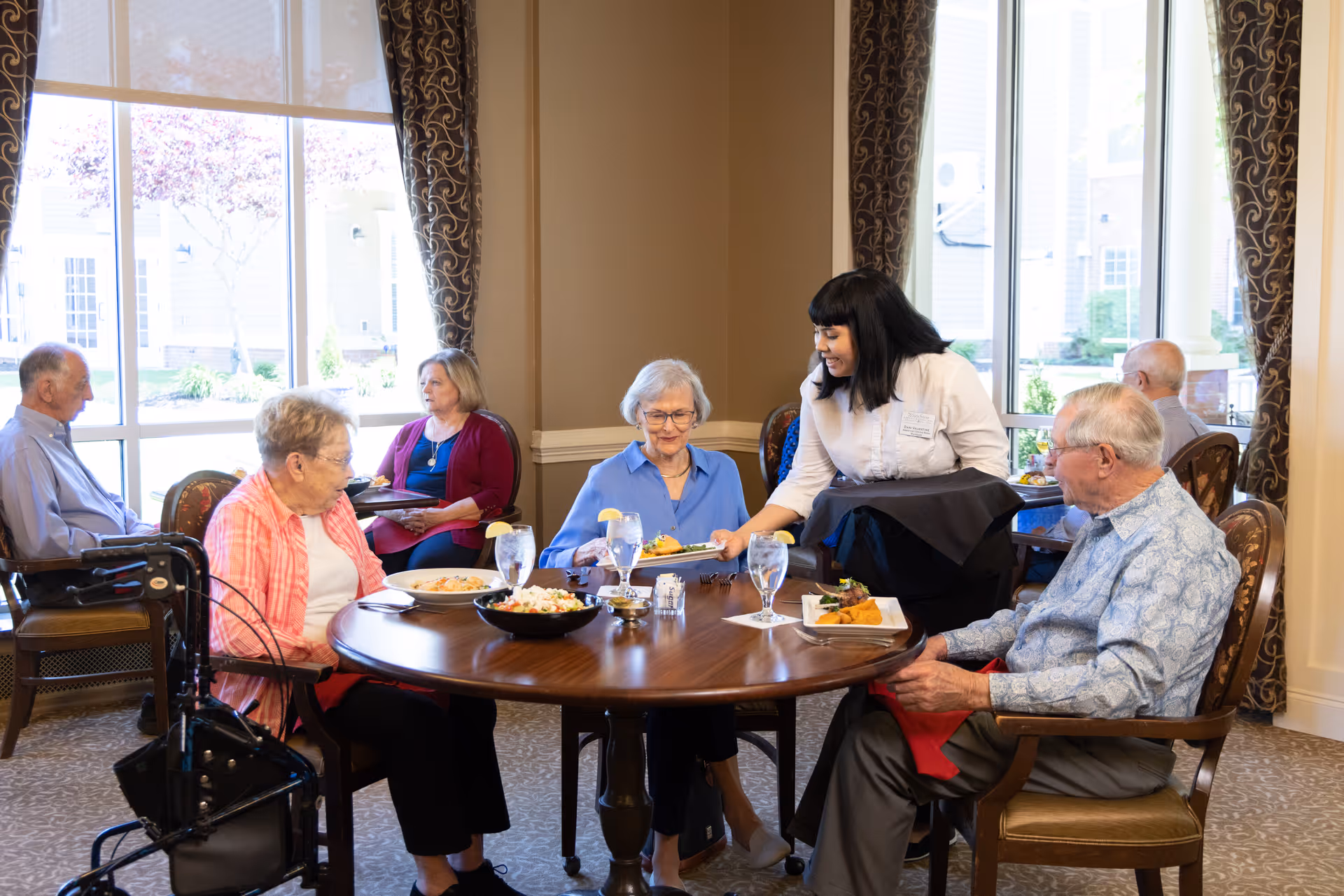 A group of elderly people seated around a dining table in a well-lit room with large windows and patterned curtains. A server is smiling and handing a plate of food to one of the elderly women. The table has plates of food, glasses of water, and a bowl of salad. Other elderly individuals are seated at tables in the background.