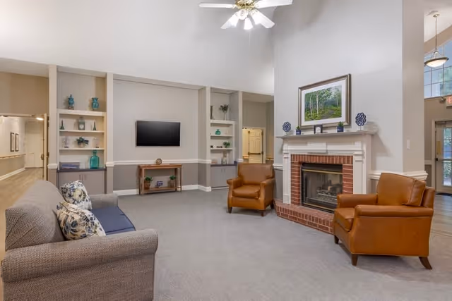 A spacious senior living facility common area with a gray sofa and two brown leather armchairs arranged around a brick fireplace with a white mantel. Above the fireplace is a framed painting of a forest scene. Built-in shelves on either side of a wall-mounted flat-screen TV display decorative items. The room has light-colored walls, a ceiling fan with lights, and a carpeted floor.