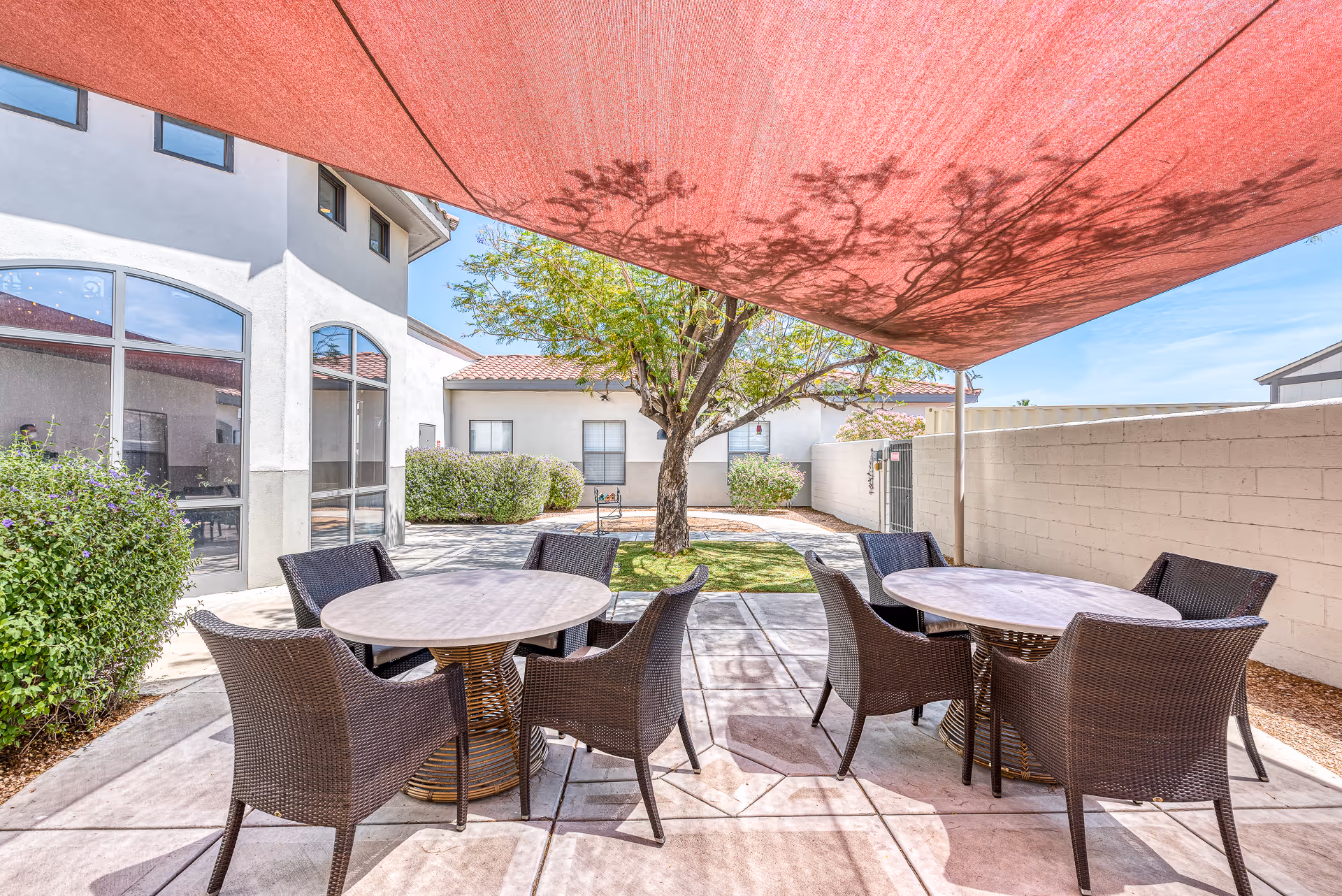 Outdoor patio area with two round tables surrounded by wicker chairs under a large red shade canopy. The patio is paved with concrete tiles and is adjacent to a building with large windows. There is a tree and some bushes in the background, along with a white brick wall enclosing the space.