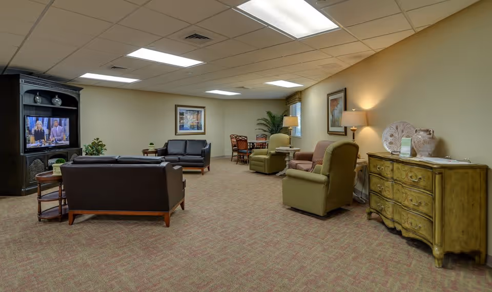 A spacious senior living lounge with sofas and armchairs arranged around a TV, a dining table in the back, and a decorative sideboard against the wall.