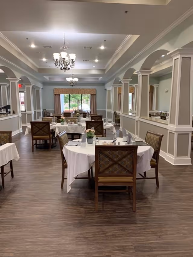 A spacious dining room with several round and rectangular tables covered with white tablecloths. Each table is set with napkins, glasses, and small flower arrangements. The room features wooden chairs with patterned upholstery, chandeliers hanging from the ceiling, large windows with curtains, and decorative columns and arches along the walls.