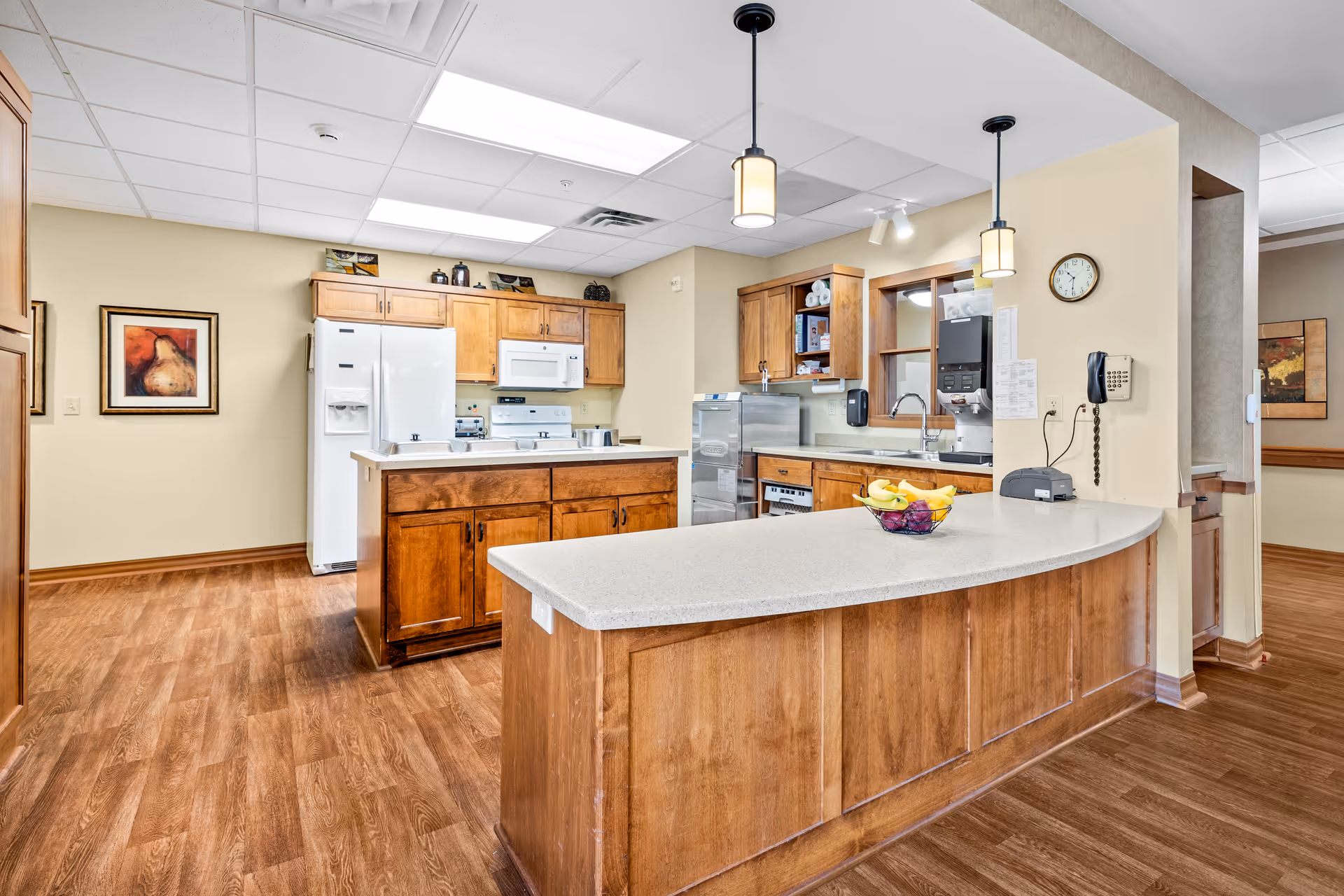 A bright, clean kitchen area with wooden cabinets and a white countertop island. The kitchen features a white refrigerator, microwave, stove, and a sink. There are two pendant lights hanging above the island, a wall clock, a telephone, and a bowl of fruit on the counter. The floor is wood, and there are framed pictures on the walls.