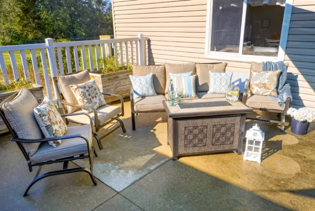 Outdoor patio area with cushioned chairs arranged around a decorative fire pit table. The patio is adjacent to a beige siding building with a window. There are decorative pillows on the chairs, a white lantern on the ground, a potted plant with white flowers, and a wooden planter box with greenery along the white railing.