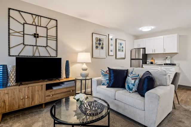 Bright living room with a gray sofa and patterned pillows, glass coffee table, TV on a wooden console, wall decor, and a small kitchenette in the background.