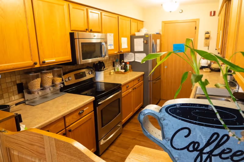 A kitchen with wooden cabinets, a stainless steel stove and microwave, a refrigerator with papers attached, cereal dispensers on the counter, and a plant in the foreground with a decorative coffee cup sign.