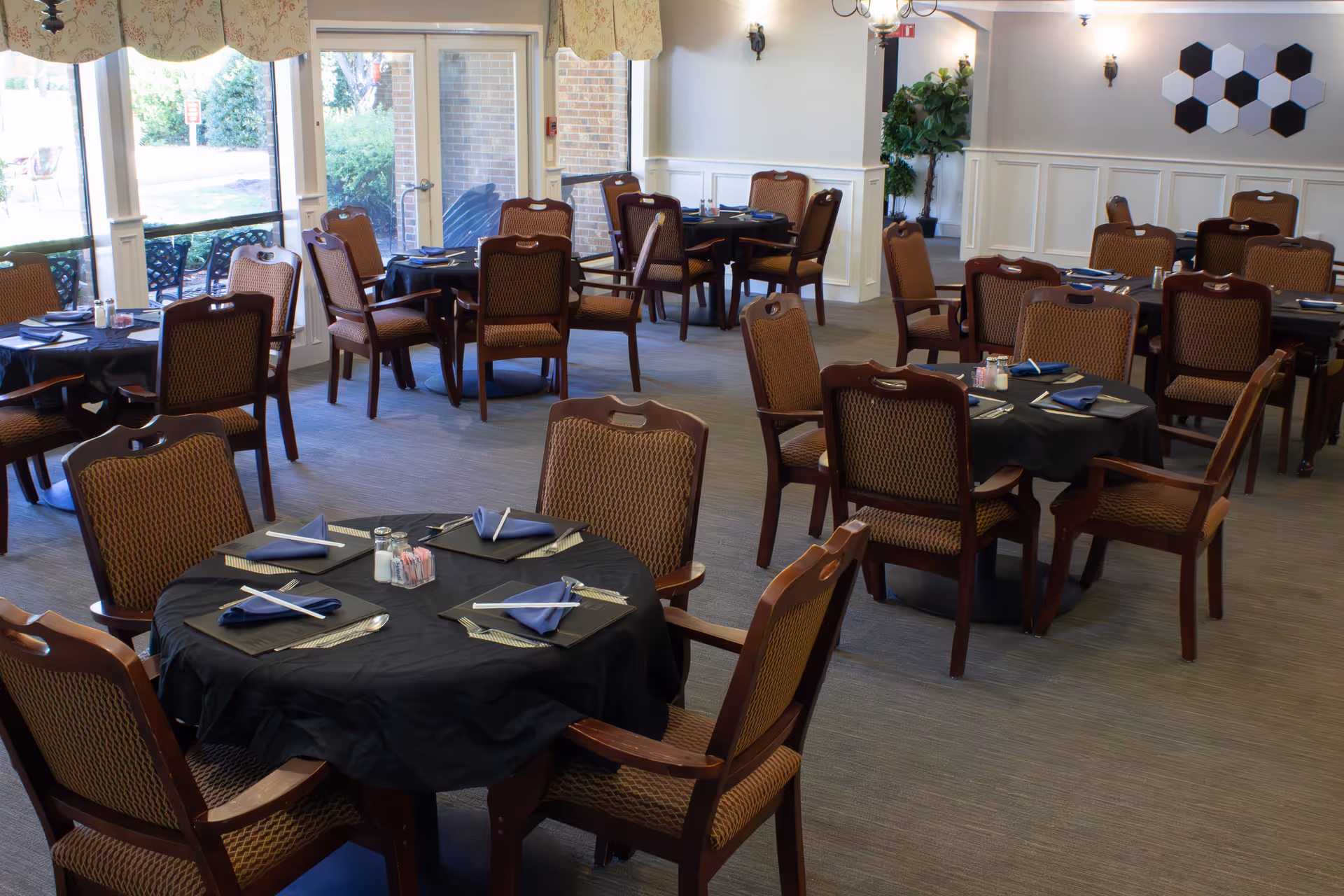 Communal dining room with round tables set with black tablecloths, blue napkins and wooden chairs by large windows.