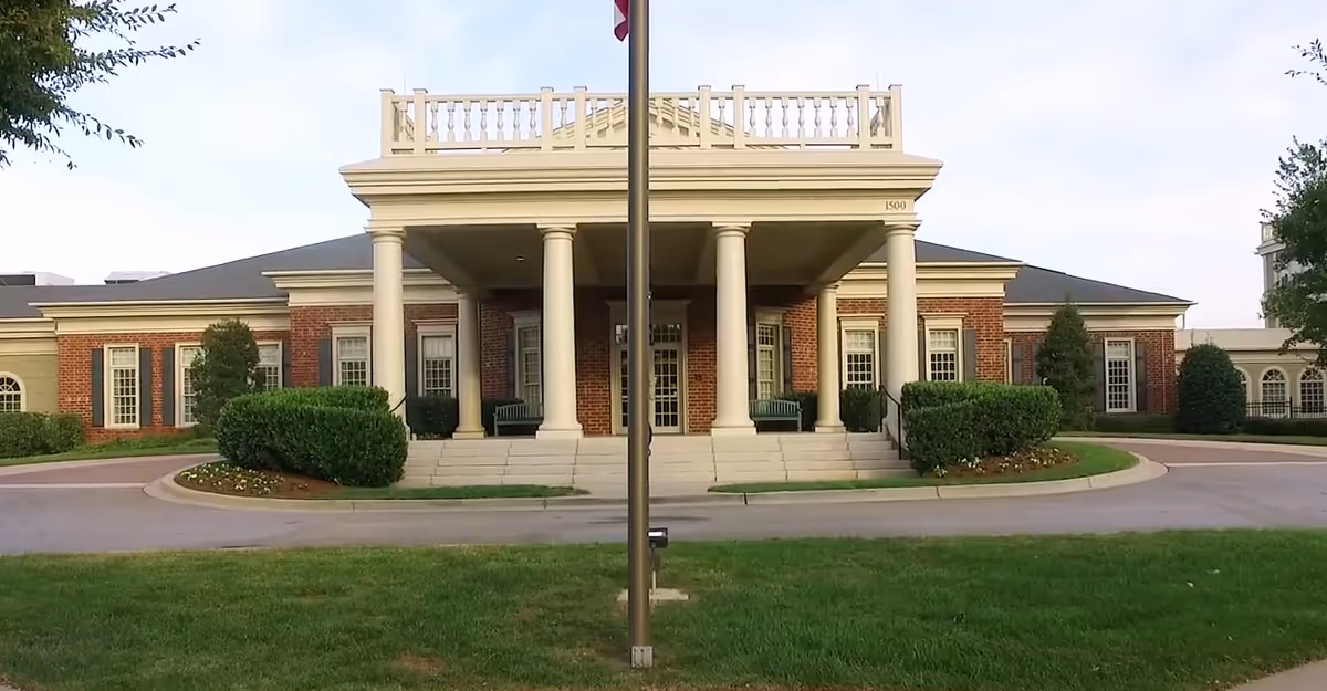 Front exterior view of a large brick building with white columns and a covered entrance, surrounded by neatly trimmed bushes and a green lawn. A flagpole stands in the center foreground.