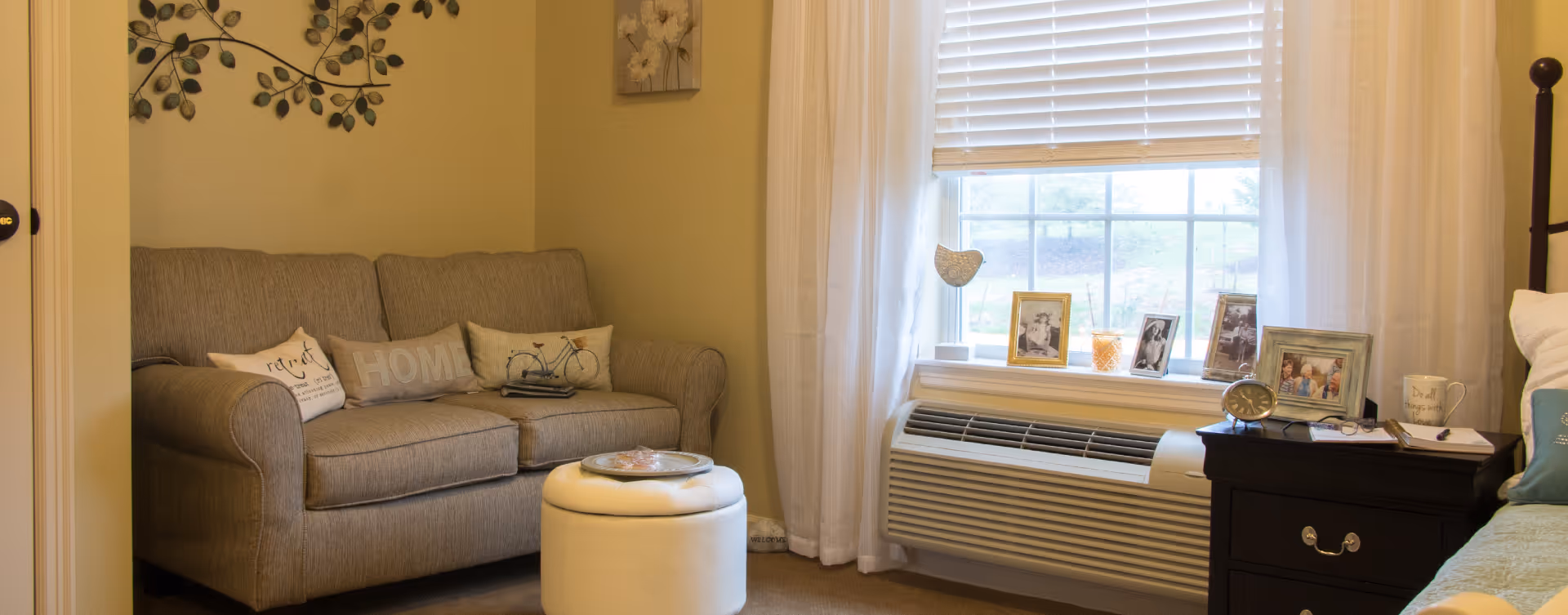 A cozy living area in a senior living facility featuring a gray loveseat with decorative pillows, a small white ottoman with a tray on top, a window with white blinds and sheer curtains, and a dark wooden nightstand with framed photos, a clock, a mug, and a notebook. The walls are painted light yellow and decorated with wall art.