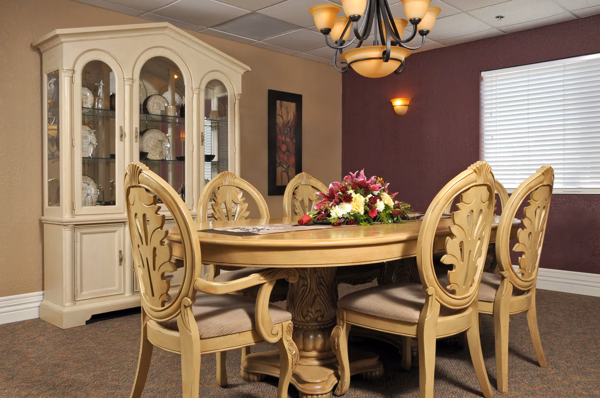 A dining room with a wooden oval table surrounded by six ornate wooden chairs with cushioned seats. A floral centerpiece decorates the table. Behind the table is a cream-colored china cabinet displaying decorative plates and glassware. The walls are painted beige and dark maroon, with a framed artwork and a window with white blinds. A chandelier with multiple lights hangs above the table.