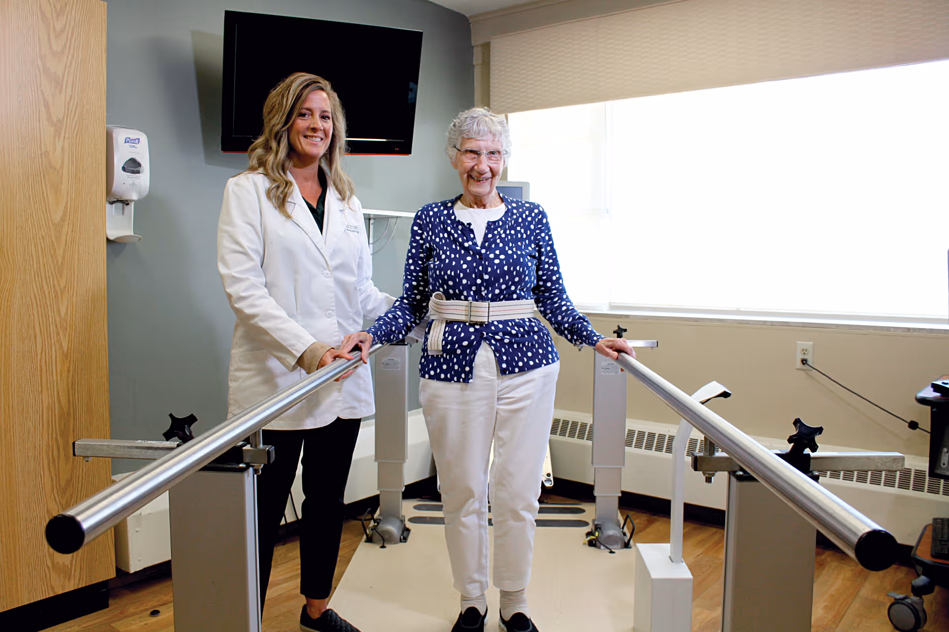 An elderly woman wearing a blue polka dot cardigan and white pants is standing between parallel bars in a rehabilitation or physical therapy room, supported by a female healthcare professional in a white coat. The room has a wooden floor, a wall-mounted TV, and a window with blinds letting in natural light.
