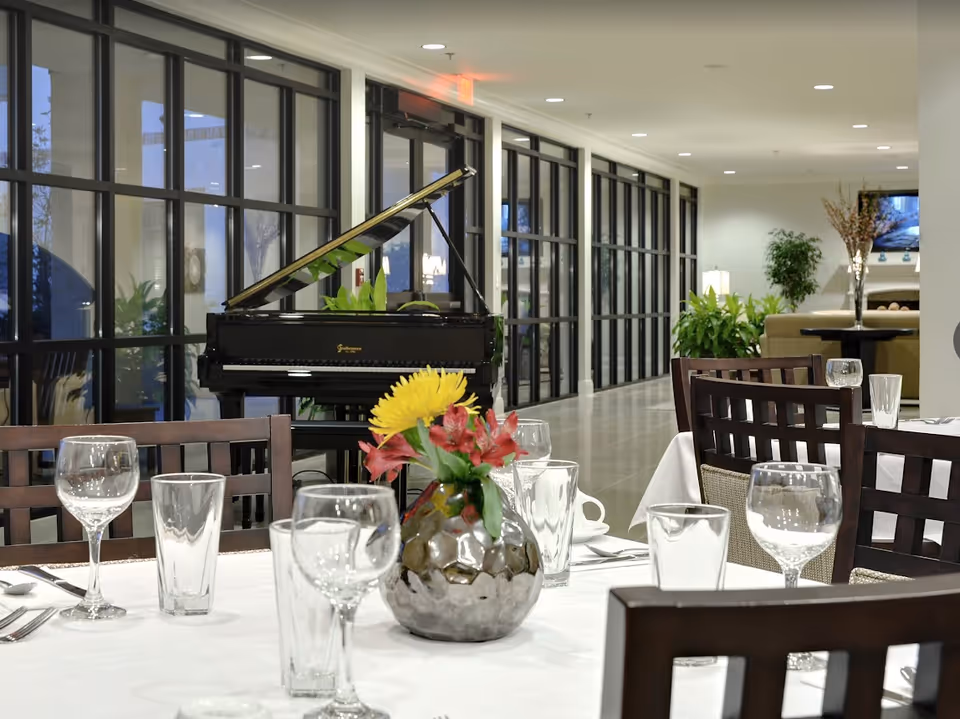 A dining area with tables set with glasses, cups, and silverware. A vase with yellow and red flowers is on the table in the foreground. In the background, there is a black grand piano and large windows along the hallway. The space is well-lit with ceiling lights and decorated with plants.