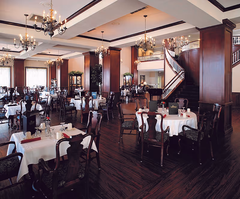 Elegant dining room with multiple round tables covered in white tablecloths, set with cups, glasses, and red napkins. The room features dark wooden chairs with patterned cushions, dark wood flooring, large wooden columns, chandeliers hanging from the ceiling, and a grand staircase in the background.