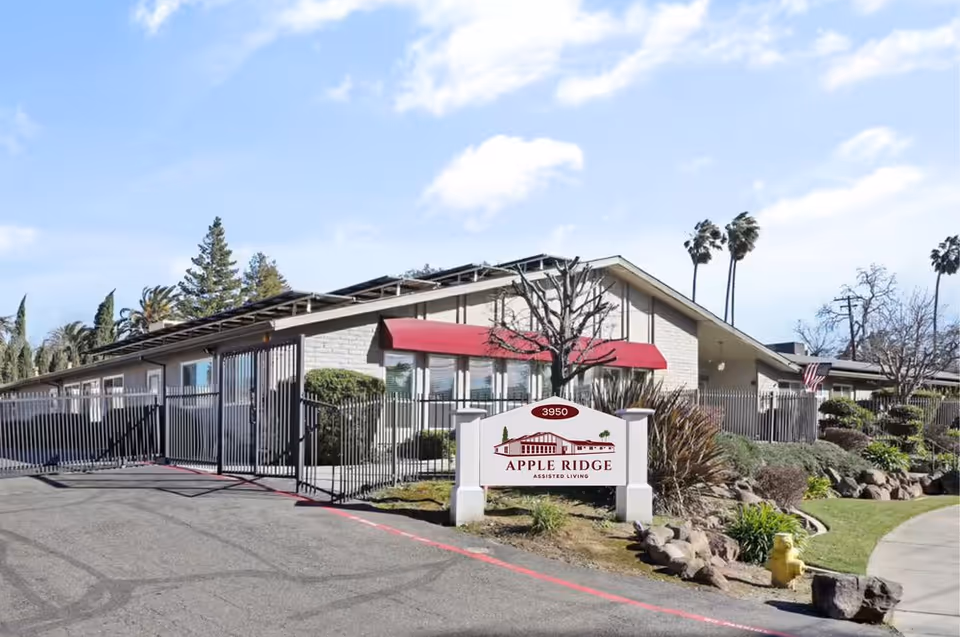 Exterior view of Apple Ridge Assisted Living facility showing a single-story building with a gated entrance, red awnings over windows, landscaping with bushes and trees, and a sign with the facility name and address 3950.