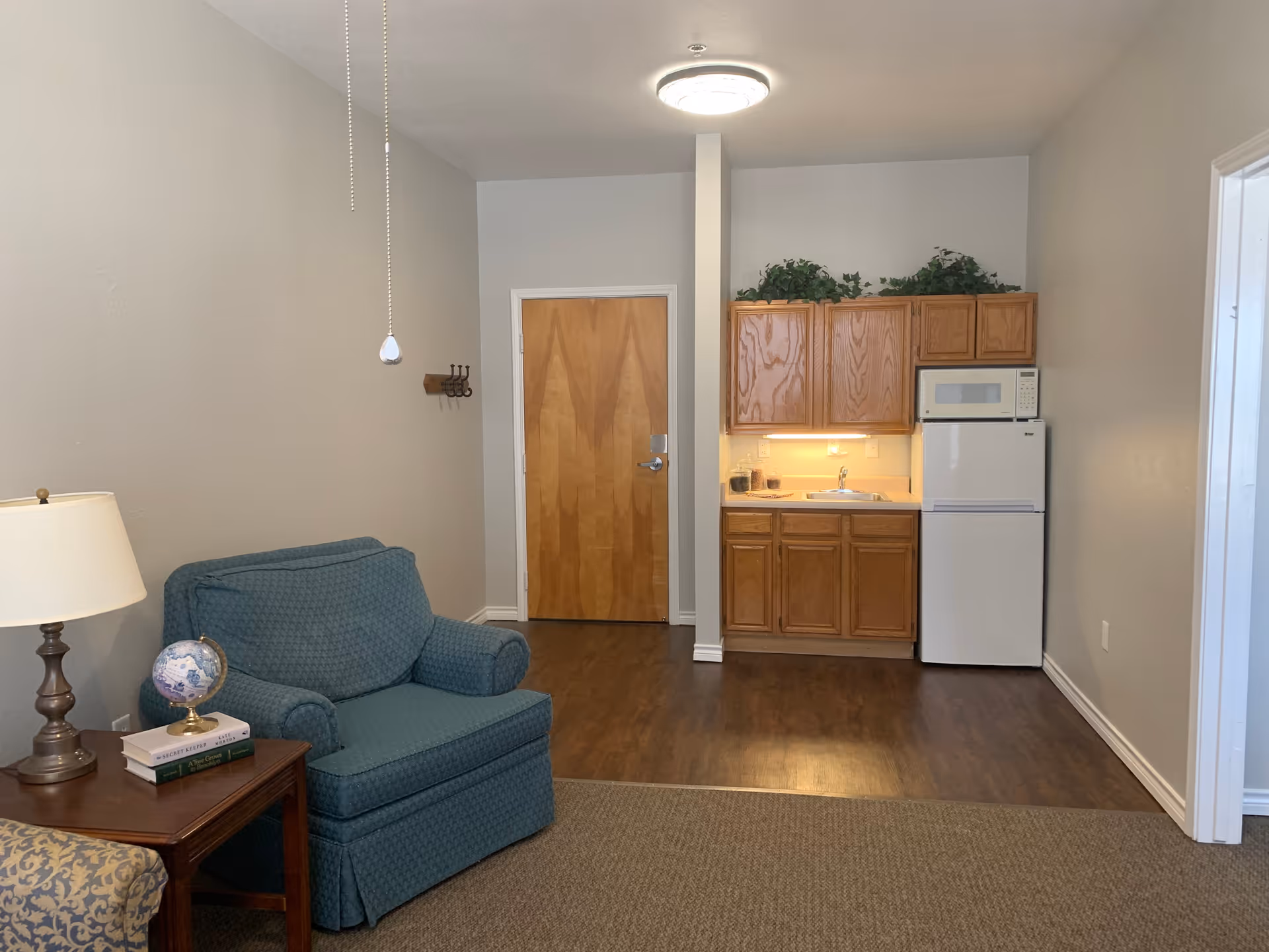 Studio-style living area with a blue upholstered armchair and side table in front of a small kitchenette with wooden cabinets, microwave, and refrigerator.