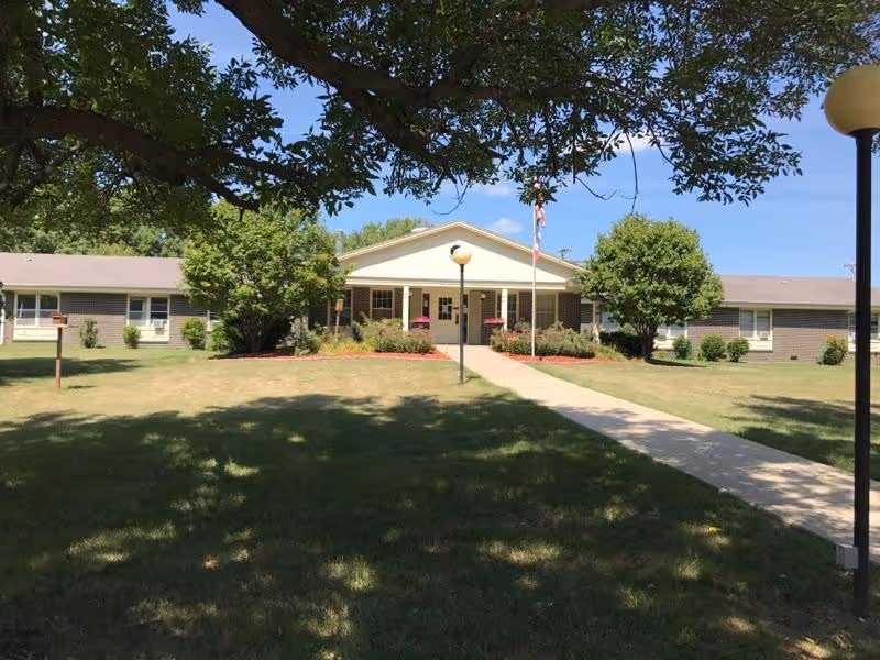 Front exterior view of a single-story brick building with a white entrance porch, surrounded by green grass, trees, and a concrete walkway leading to the entrance. There are two lamp posts and an American flag near the entrance under a clear blue sky.