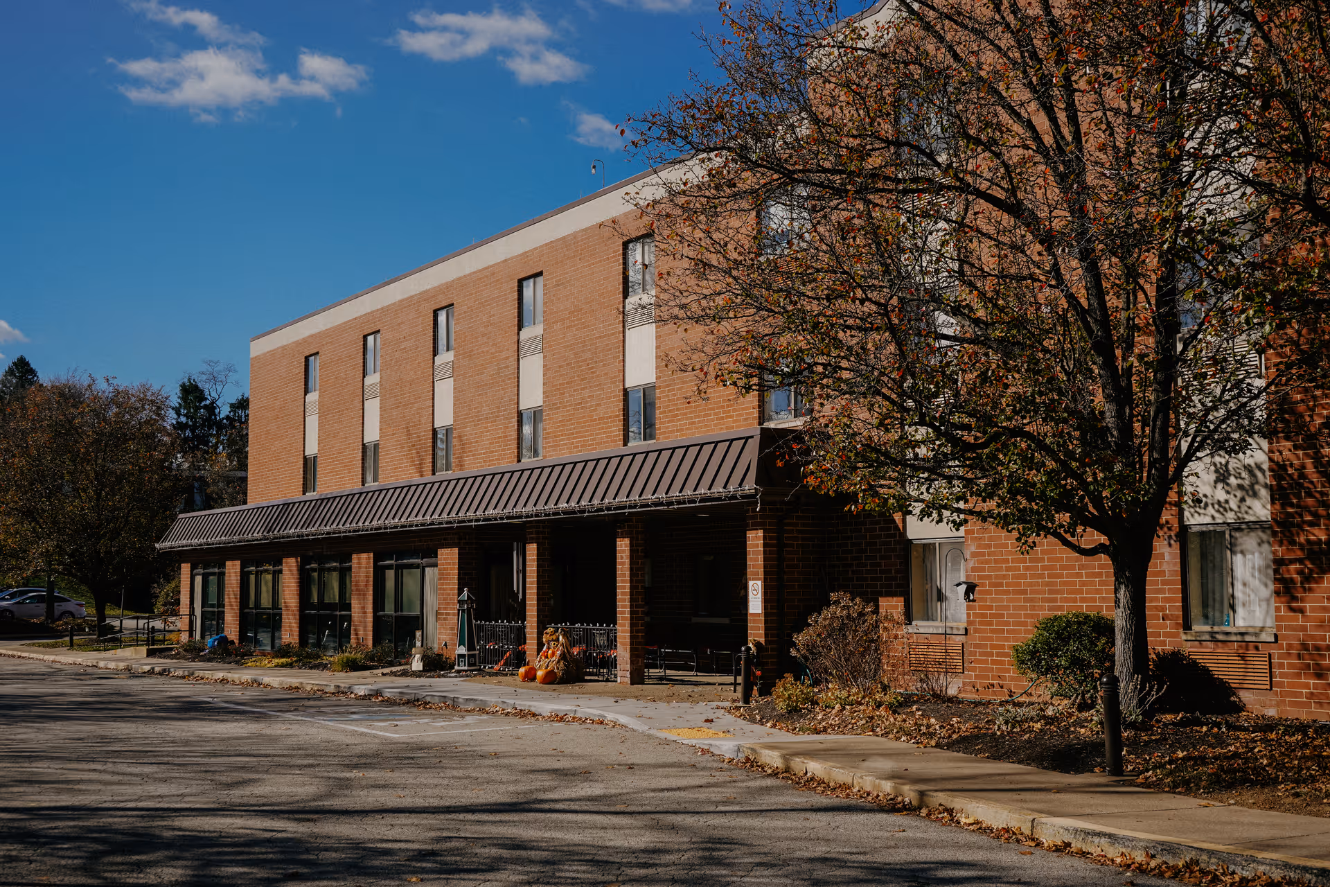 Exterior view of a three-story brick building with large windows on the ground floor and smaller windows on the upper floors. There is a covered entrance area with columns and some autumn decorations including pumpkins. Trees with fall foliage are visible around the building under a clear blue sky.