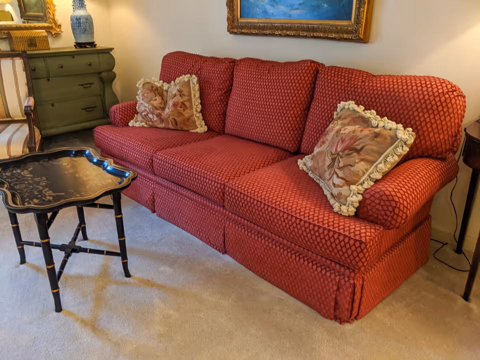 A cozy living room area featuring a red patterned sofa with two decorative floral pillows. In front of the sofa is a small black wooden table with intricate designs. To the left, there is a green chest of drawers with a blue and white vase on top, and a striped armchair partially visible. A framed painting hangs on the wall above the sofa.