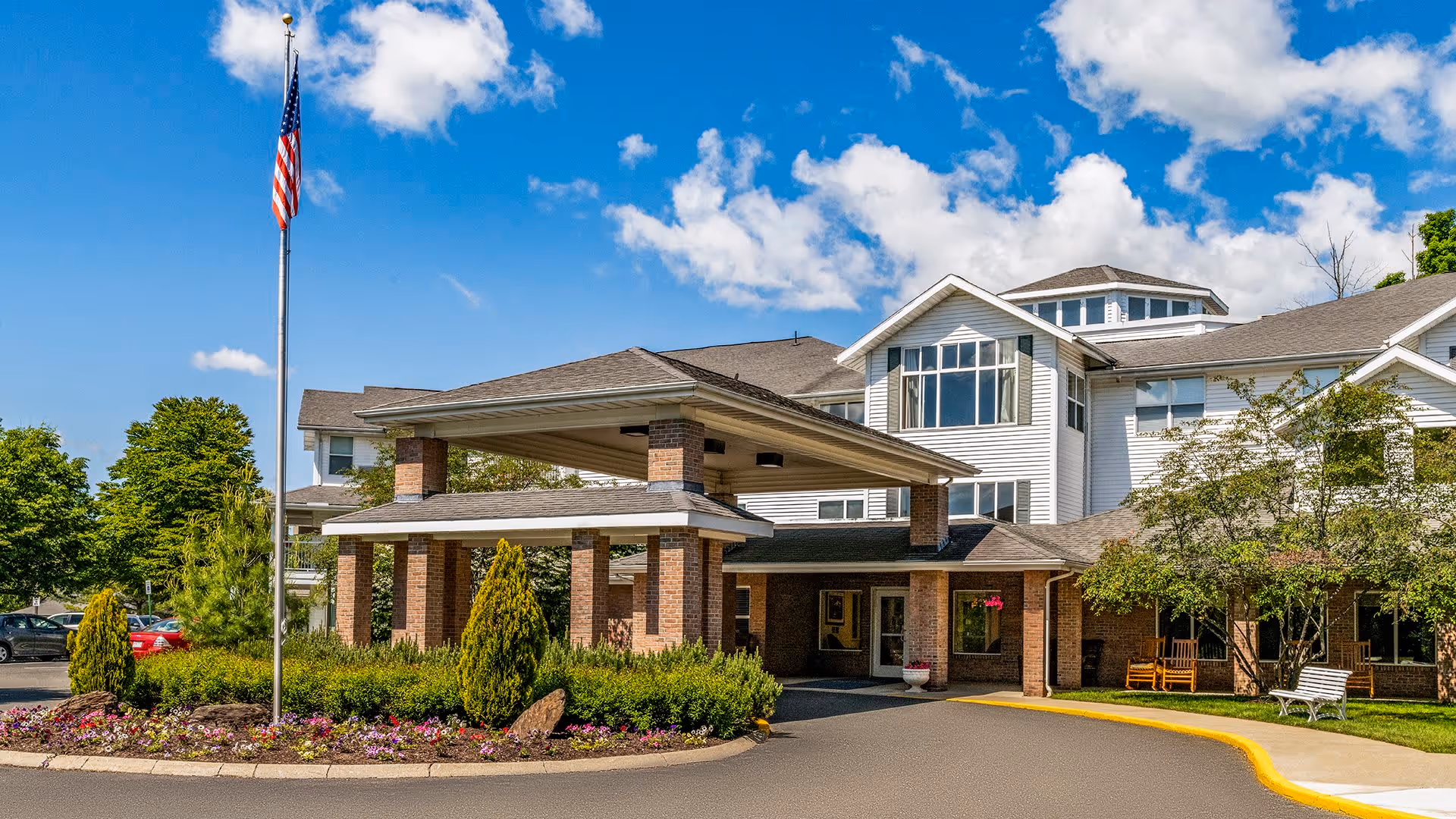 Exterior view of Holiday Devonshire Estates, a senior living facility with a covered entrance supported by brick pillars, landscaped flower beds, an American flag on a flagpole, and a bright blue sky with scattered clouds.