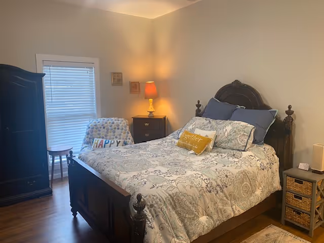 A cozy bedroom with a dark wooden bed, nightstands, a lamp, an armoire, and a window with blinds.