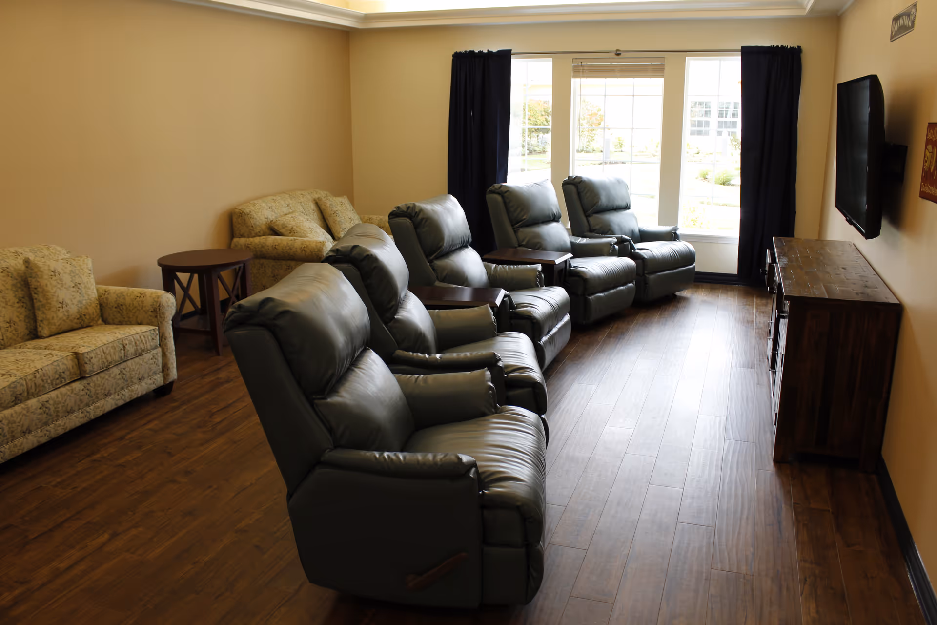 A living room with four dark leather recliners arranged in a row facing a wall-mounted flat-screen TV. There are two floral-patterned sofas along the left wall, a wooden side table between them, and a large window with black curtains letting in natural light. The floor is wooden and the walls are painted beige.