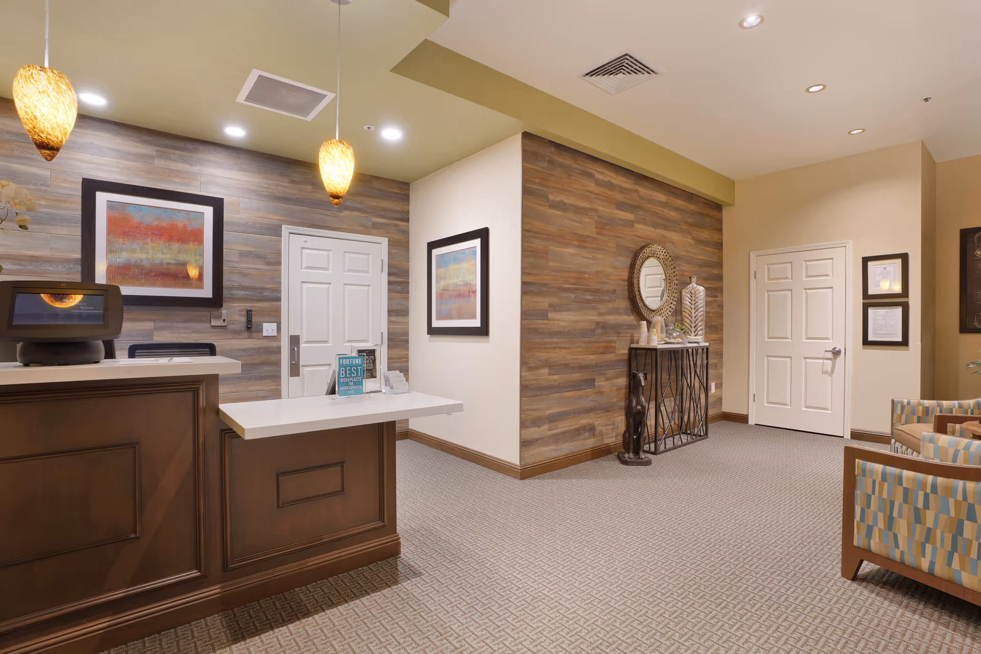 Reception area of MuirWoods Memory Care featuring a wooden front desk with a computer, two hanging pendant lights, framed abstract artwork on wood-paneled walls, a decorative console table with a round mirror, and patterned armchairs on a carpeted floor.